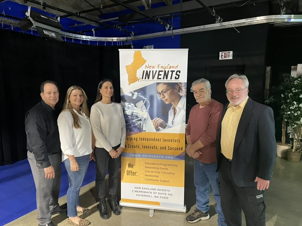 Five people stand indoors next to a New England Invents banner, smiling at the camera. The banner highlights inventor support services. The group stands on a black and blue stage with studio lighting overhead.