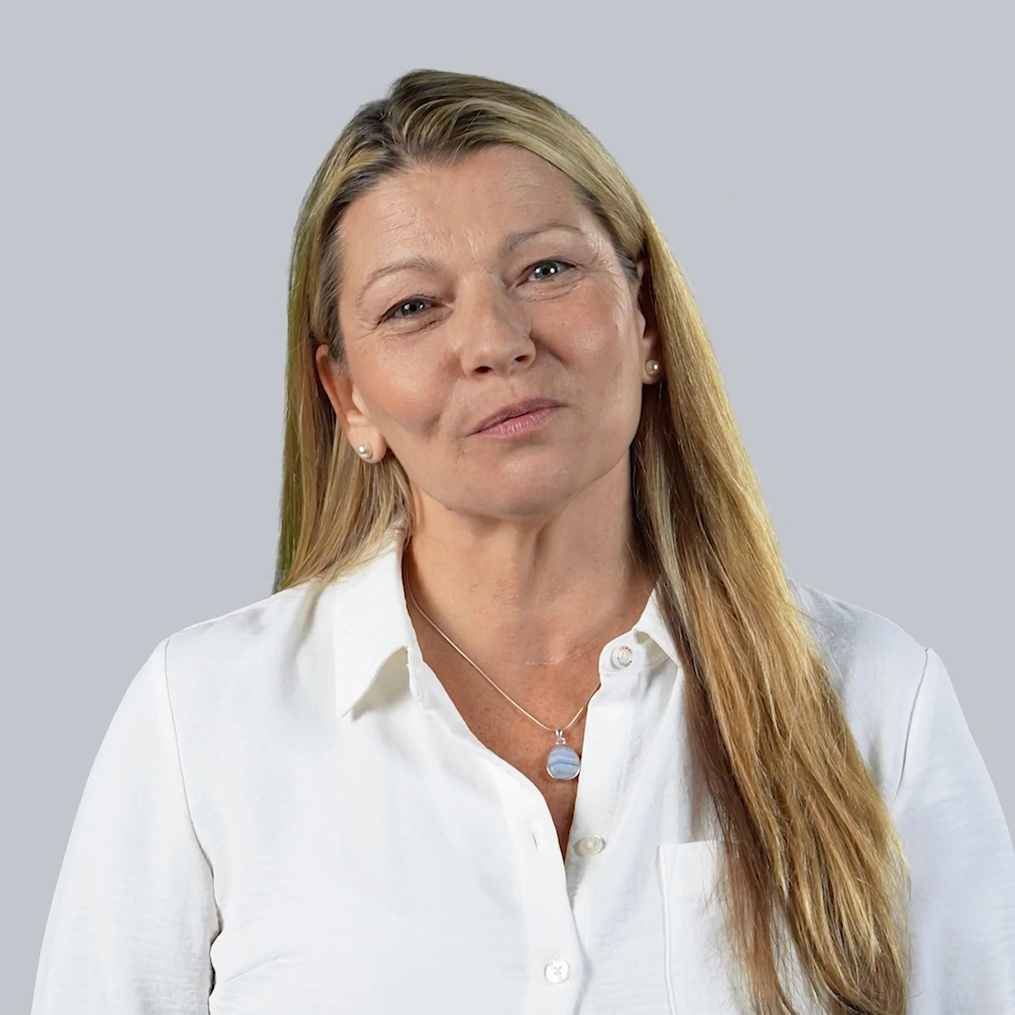 A woman with long blonde hair wearing a white blouse and a pendant necklace stands in front of a plain light gray background, smiling softly at the camera.